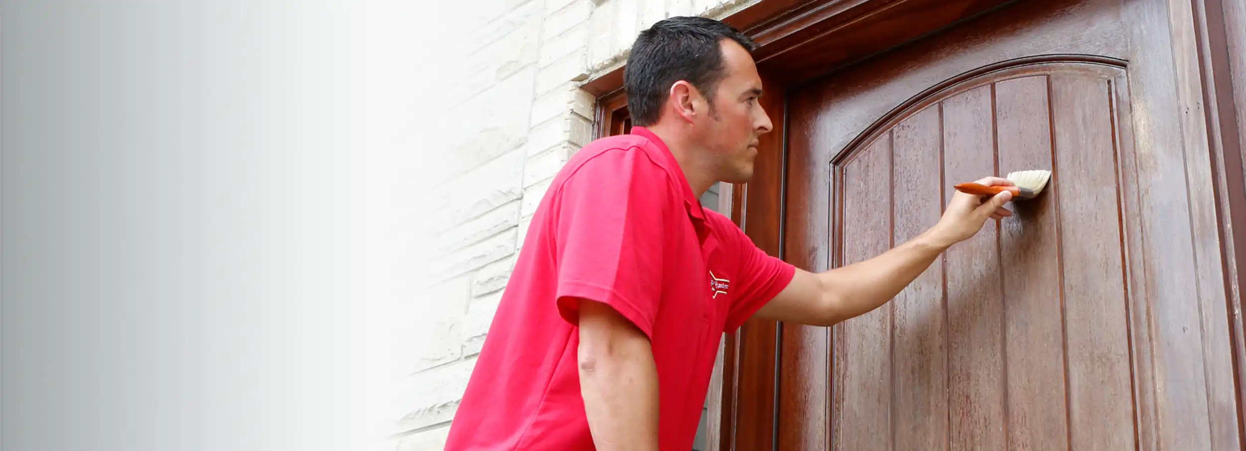 Mr. Handyman technician in red polo shirt staining a wood front door with a paintbrush.