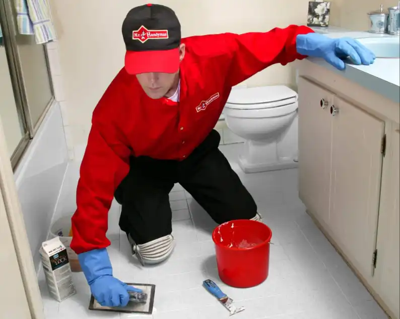 A handyman from Mr. Handyman using a trowel to apply a layer of sealant on a tiled bathroom floor after flooring repairs have been completed.