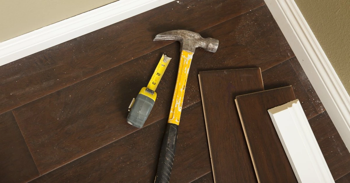 A hammer, tape measure, laminate floorboards and new baseboards on the floor of a home where flooring installation is being completed.