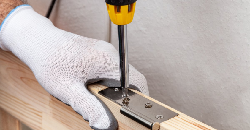 A handyman wearing work gloves and using a screwdriver to attach a new hinge to the side of a door during an appointment for door repair in Wichita, KS.