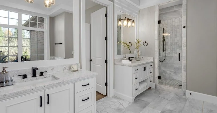 A modern bathroom after a bathroom remodeling project has been completed to install two separate vanities and a door between them, as well as grey, marble tile, and a glass shower door.