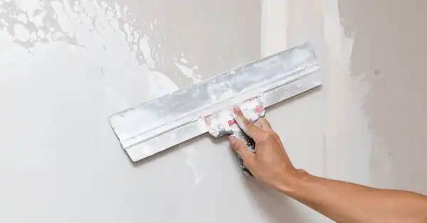 A handyman’s hand holding a wide putty scraper as they use it to smooth out a layer of joint compound during an appointment for drywall repair in Charleston, SC.
