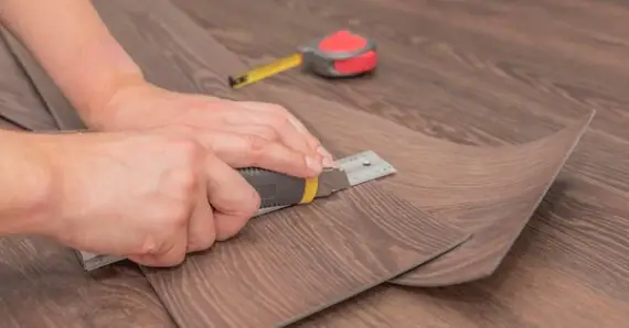 A handyman using a utility knife, ruler, and tape measure to cut pieces of vinyl flooring during an appointment for flooring repair in Tulsa, OK.