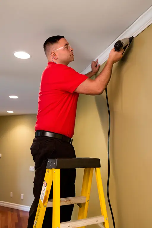 A handyman from Mr. Handyman standing on a ladder and using a nail gun to install crown molding on the ceiling of a home.