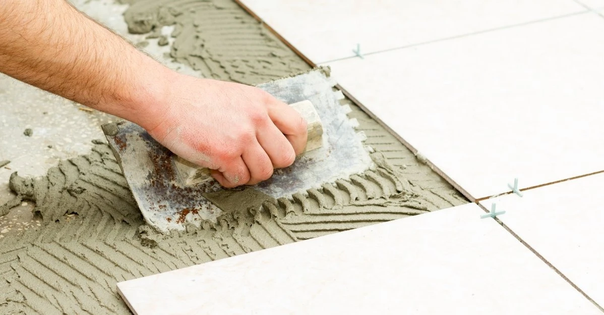 A handyman using a notched trowel to lay a layer of grout across a subfloor before laying a new piece of tile during an appointment for tile installation in Denton, TX.