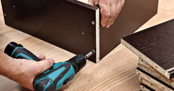 A handyman using a power drill to attach two wooden boards during a furniture assembly project.