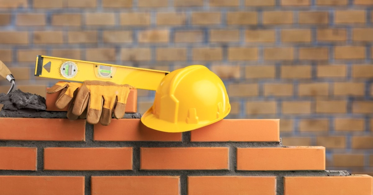 A half-finished brick wall with a new brick, a trowel covered in mortar, a level, a pair of work gloves and a hard hat set aside by a handyman providing services for masonry in Dallas, TX.