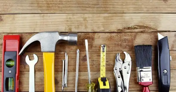 Tools like a hammer, paintbrush, tape measure, and wrench laid out in a straight line on a wood surface.