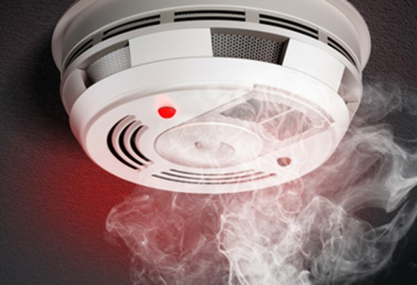 A smoke detector installed on the ceiling of a home to reduce aging in place challenges, with a red light shining in the presence of smoke swirling up around the area of the smoke detector.