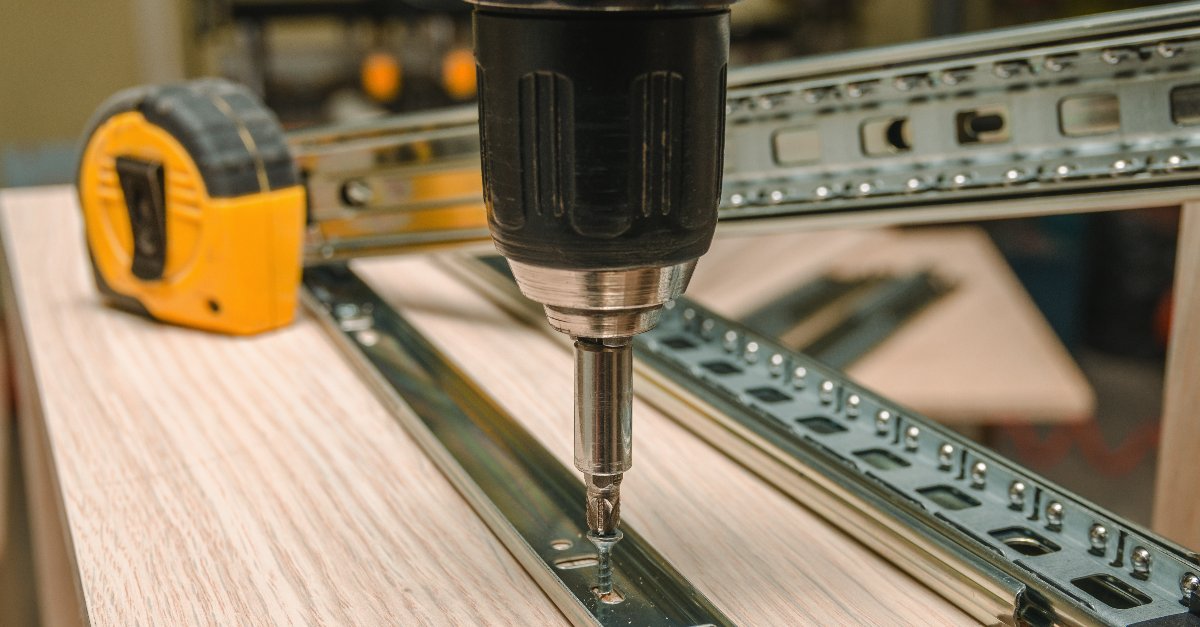 A close up of a handyman using a drill to attach tracks on the side of a piece of furniture while providing furniture assembly service.