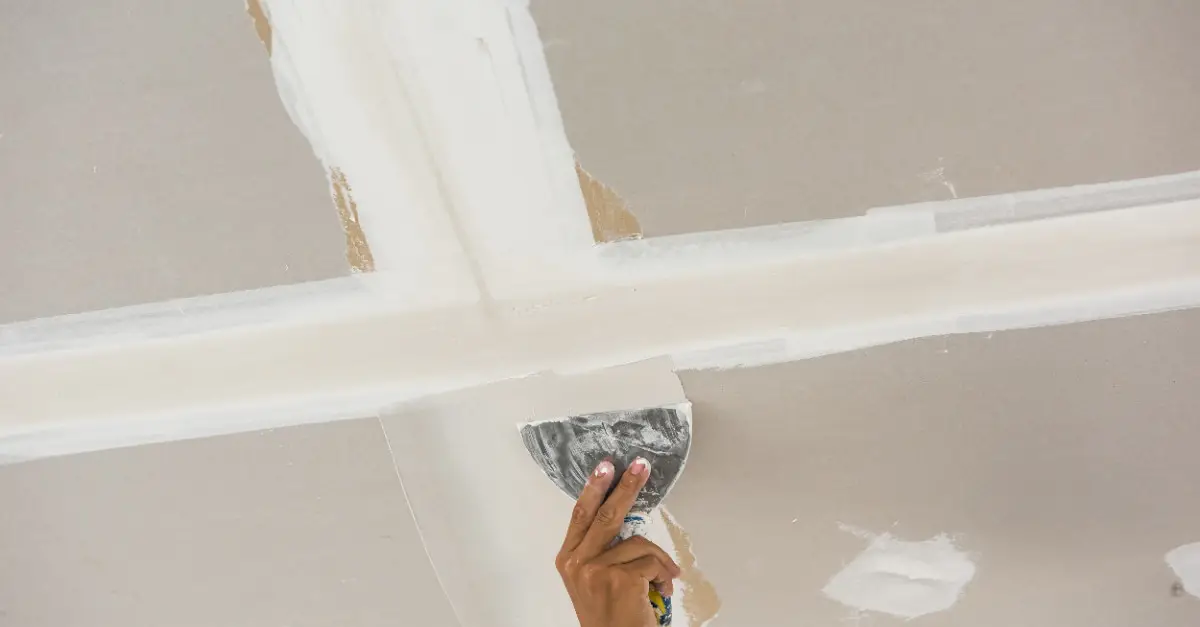  A handyman using a putty knife to spread joint compound over the seam tape covering the joint between two pieces of drywall on a residential ceiling.