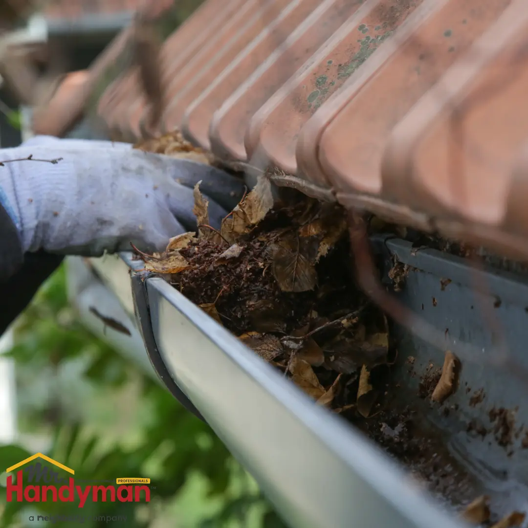 A gloved hand clearing out a clogged gutter filled with dry leaves and debris.