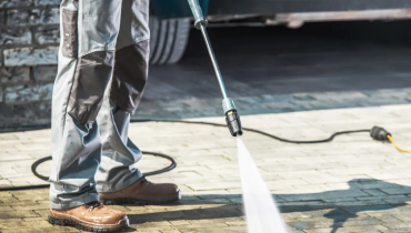 A worker using a pressure washer to clean a paved driveway, with the high-pressure water stream directed at the surface.