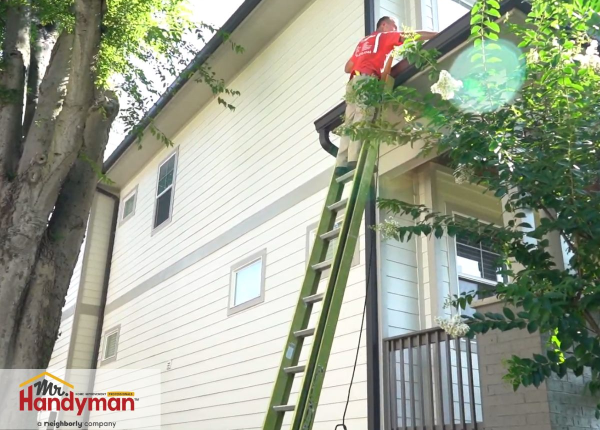Mr. Handyman technician cleaning house gutters on a ladder during a professional home maintenance service.