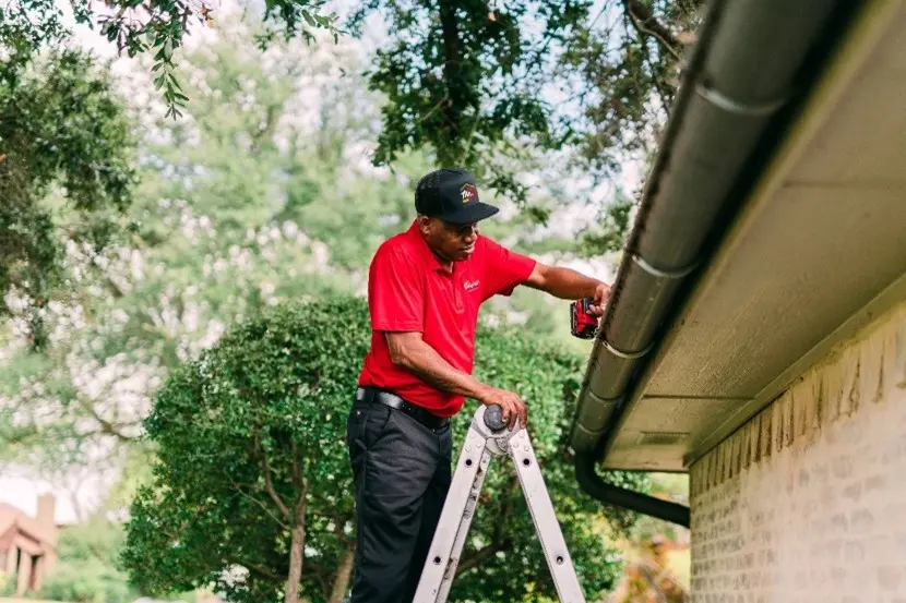 A Mr. Handyman technician standing on a ladder and cleaning a gutter.