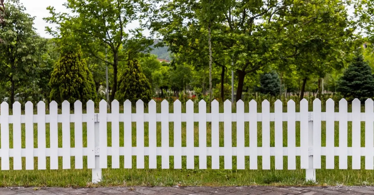A white picket fence installed where a paved stone area meets a large patch of grass and trees.